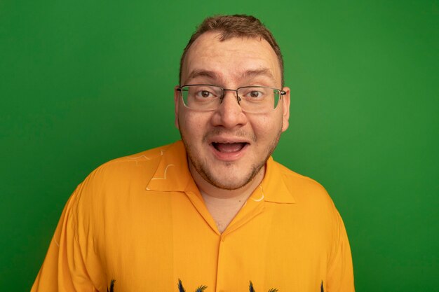 Hombre feliz con gafas y camisa naranja sorprendido sonriendo de pie sobre la pared verde