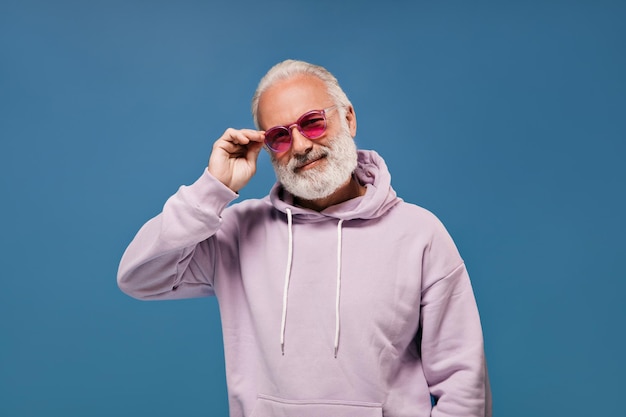 Hombre feliz con gafas de sol rosas posando sobre fondo azul Chico elegante con barba gris con capucha lila claro mirando a la cámara