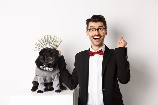Hombre feliz ganando dinero, disfrazado y mostrando dólares cerca de su lindo perro negro en traje, de pie sobre fondo blanco.