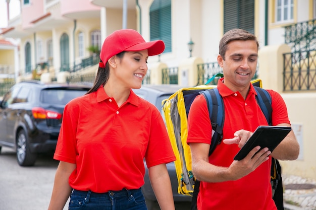 Hombre feliz con gorra roja que muestra la dirección a su colega. Mensajeros sonrientes trabajando juntos y entregando pedidos a pie.