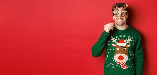 Foto gratuita hombre feliz guapo en suéter de navidad sosteniendo máscara de fiesta y sonriendo disfrutando de celebración de año nuevo ...