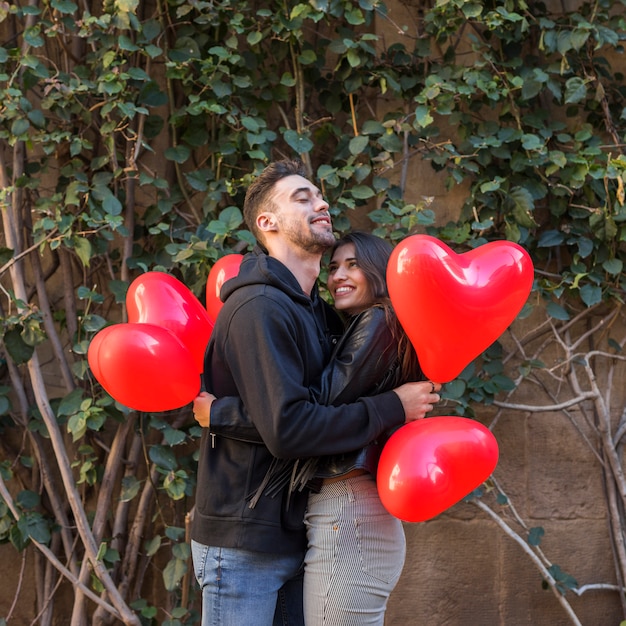 Hombre feliz joven que abraza a la mujer sonriente y que sostiene los globos en la forma de corazones