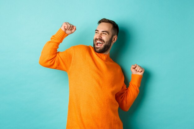 Hombre feliz levantando el brazo, sintiéndose como un ganador, triunfando y celebrando, de pie sobre una pared turquesa clara.