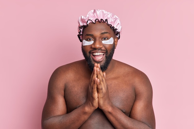El hombre feliz mantiene las palmas juntas tiene buen humor aplica parches humectantes debajo de los ojos usa soportes de gorro de ducha con el cuerpo desnudo aislado sobre una pared rosa