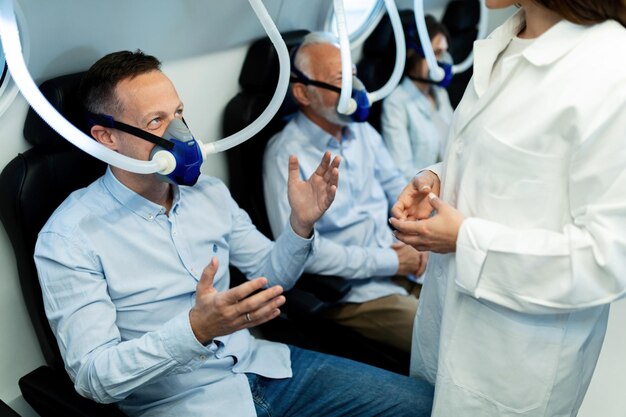 Hombre feliz con máscara de oxígeno y hablando con una doctora en cámara hiperbárica en la clínica