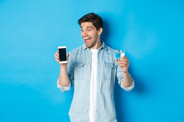 Hombre feliz mirando emocionado en la pantalla del teléfono móvil, sosteniendo un vaso de agua, de pie sobre fondo azul.