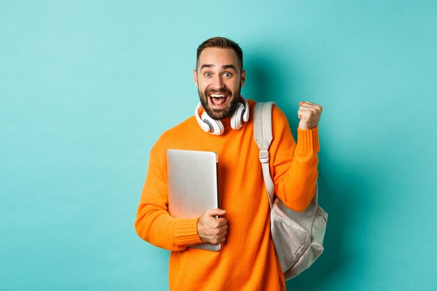 Hombre feliz con mochila y auriculares, sosteniendo el portátil y sonriendo, animando a ganar, triunfando