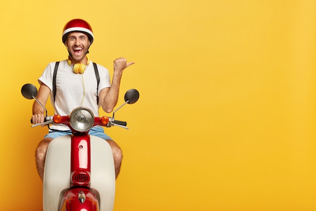 Hombre feliz monta un scooter, muestra la dirección de distancia, señala con el pulgar hacia la derecha en el espacio en blanco sobre fondo amarillo, vestido con ropa casual y casco, usa auriculares, tiene una expresión de cara alegre