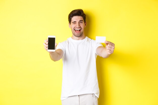 Hombre feliz mostrando una buena oferta en línea en la pantalla del teléfono móvil, sosteniendo la tarjeta de crédito y guiñando un ojo, de pie sobre un fondo amarillo.
