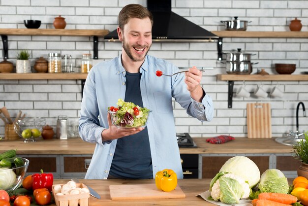 Hombre feliz de pie en la cocina comiendo ensalada fresca con tenedor