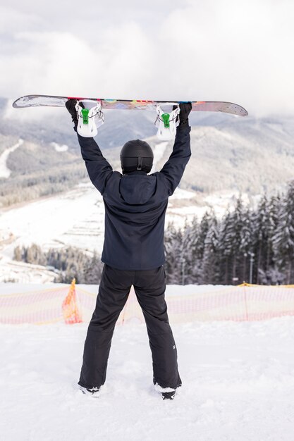Hombre feliz de pie con snowboard sobre su cabeza en la cima de la colina de las montañas