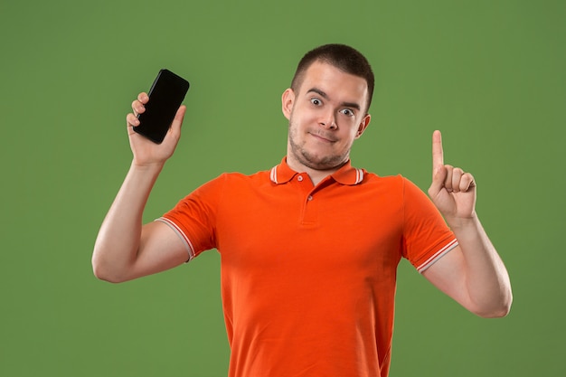 El hombre feliz que muestra en la pantalla vacía del teléfono móvil contra la pared verde.