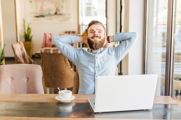 Hombre feliz que se relaja en el café con la taza de café y el ordenador portátil en el escritorio