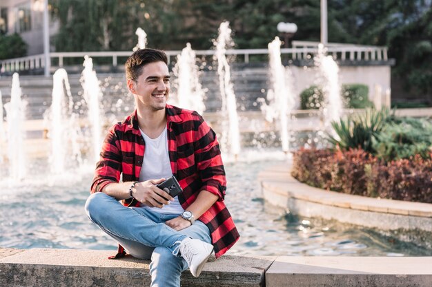 Hombre feliz que se sienta delante de la fuente que sostiene el teléfono móvil