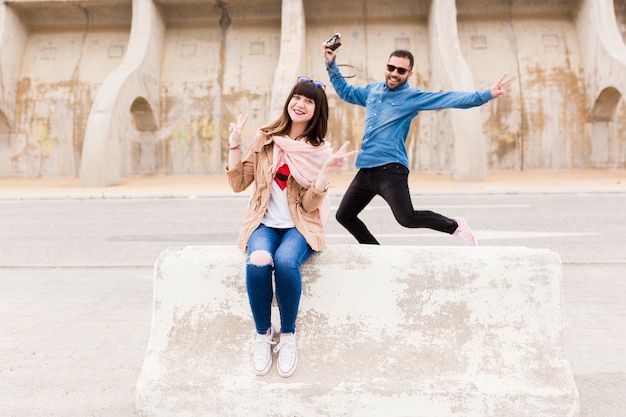 Hombre feliz que sostiene la cámara que salta delante de la mujer que se sienta sobre banco