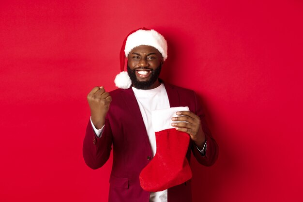 Hombre feliz regocijándose, recibiendo regalos en calcetín de Navidad, haciendo bomba de puño y sonriendo satisfecho, de pie sobre fondo rojo.