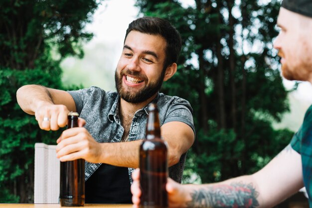 Hombre feliz sentado con amigo abriendo la botella de cerveza al aire libre