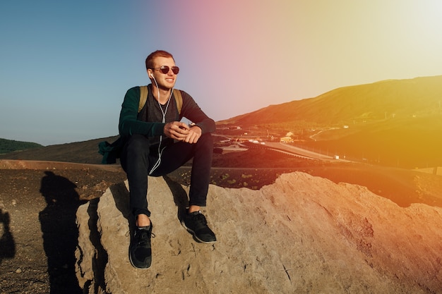 Hombre feliz sentado en el rock y escuchando música en auriculares al atardecer