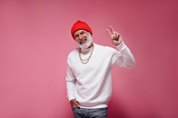 Hombre feliz con sombrero naranja y suéter blanco que muestra el signo de la paz sobre fondo rosa Chico guapo adulto con ropa moderna sonriendo