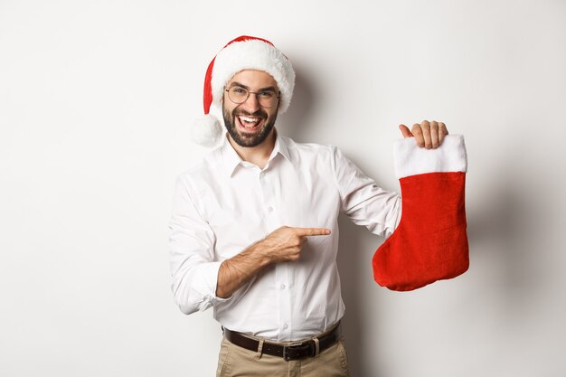 Hombre feliz con sombrero de santa celebrando las vacaciones de invierno, apuntando al calcetín de Navidad y sonriendo, sosteniendo regalos, de pie
