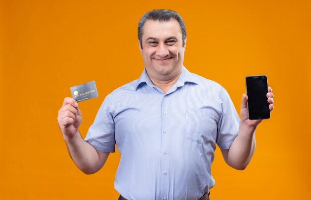 Hombre feliz y sonriente en camisa azul a rayas verticales mostrando tarjeta de crédito y teléfono móvil mientras está de pie