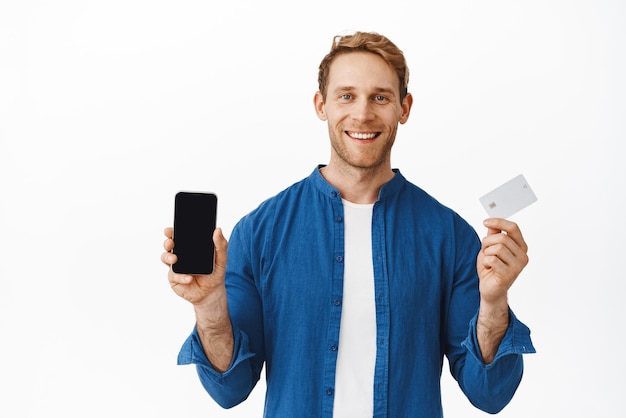 Hombre feliz sonriente que muestra la pantalla en blanco del teléfono móvil y la tarjeta de crédito bancaria recomendando la aplicación de compras en línea de la aplicación de teléfono inteligente de pie sobre fondo blanco