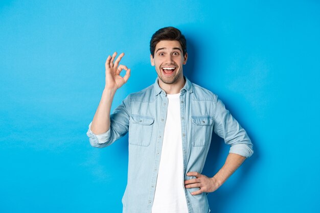 Hombre feliz sonriente que muestra el signo de ok y parece complacido, aprobando algo bueno, de pie contra el fondo azul.