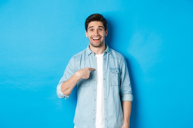 Hombre feliz y sorprendido apuntando a sí mismo, sonriendo complacido, de pie contra el fondo azul.