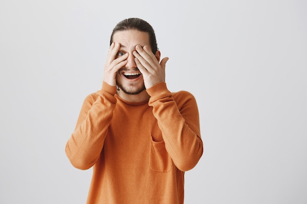 Hombre feliz sorprendido sonriendo con los ojos cerrados, mirando a través de los dedos emocionados