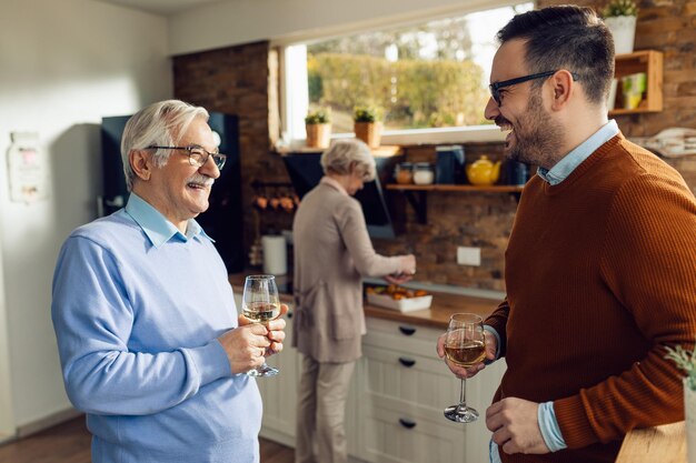 Hombre feliz y su padre mayor bebiendo vino y hablando en la cocina Mujer mayor está preparando comida en el fondo