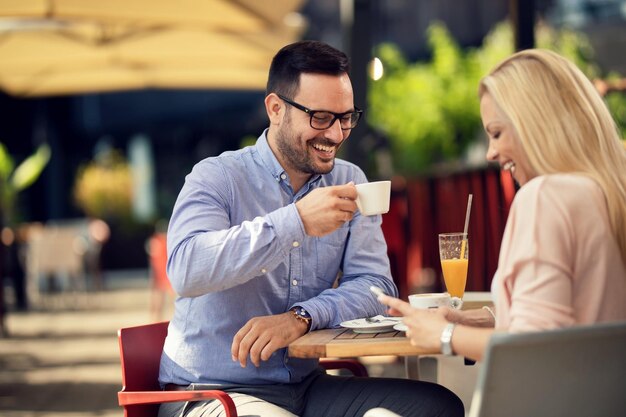 Hombre feliz tomando café y divirtiéndose con su novia mientras ella usa el celular en un café