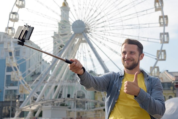 Hombre feliz tomando selfie frente a la noria y mostrando el pulgar hacia arriba gesto