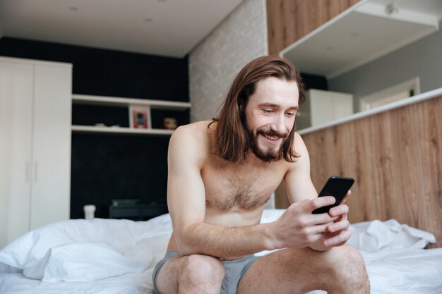 Hombre feliz usando teléfono celular sentado en la cama en el dormitorio