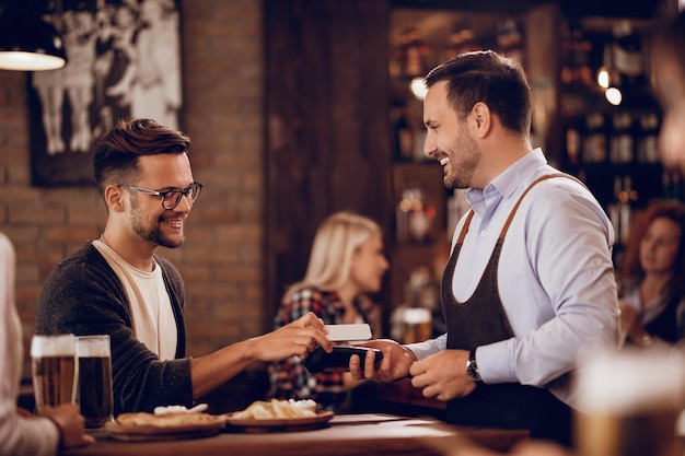 Hombre feliz usando un teléfono inteligente mientras realiza un pago sin contacto a un camarero en un bar