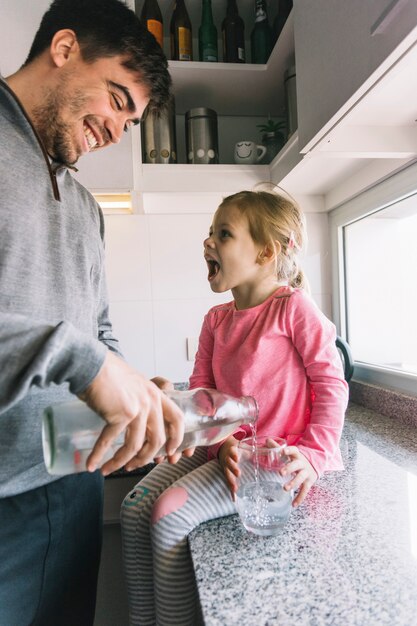 Hombre feliz vertiendo agua en vidrio mientras niña sentada en el mostrador de la cocina