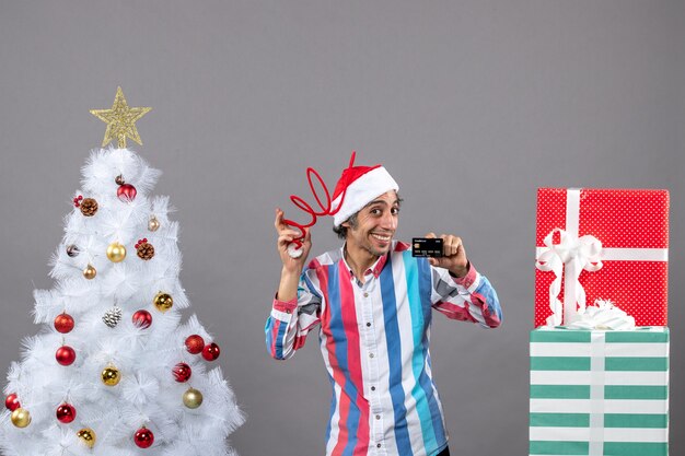 Hombre feliz de vista frontal sosteniendo su gorro de Papá Noel y tarjeta de crédito de pie cerca del árbol de Navidad blanco