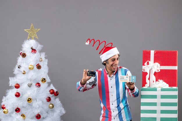 Hombre feliz de vista frontal con tarjeta de tenencia de ojo parpadeado y boleto de viaje alrededor del árbol de Navidad y regalos