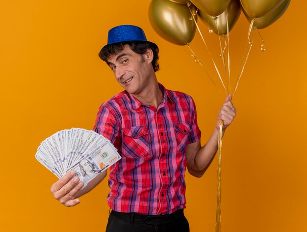 Hombre de fiesta caucásico de mediana edad complacido con sombrero de fiesta sosteniendo globos y estirando dinero hacia la cámara mirando a cámara aislada sobre fondo naranja