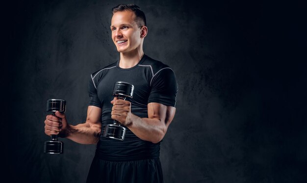 Un hombre de fitness atlético vestido con ropa deportiva sostiene un juego de pesas sobre fondo gris.