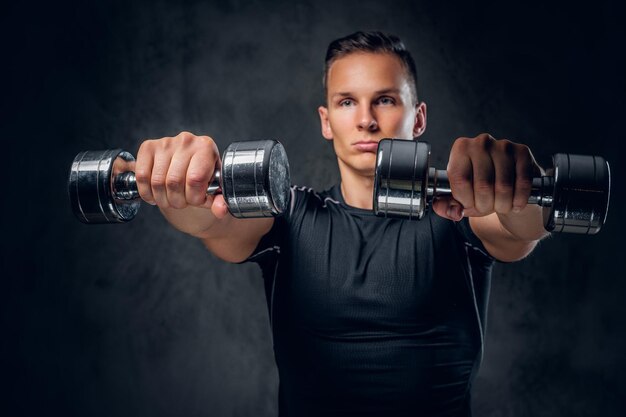 Un hombre de fitness atlético vestido con ropa deportiva sostiene un juego de pesas sobre fondo gris.