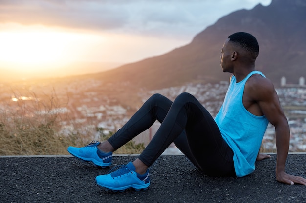 El hombre de fitness se sienta de lado, tiene piel negra, manos musculosas, vestido con ropa deportiva, mira atentamente al amanecer, posa sobre las montañas, toma un descanso después de una carrera intensa. Deporte, naturaleza