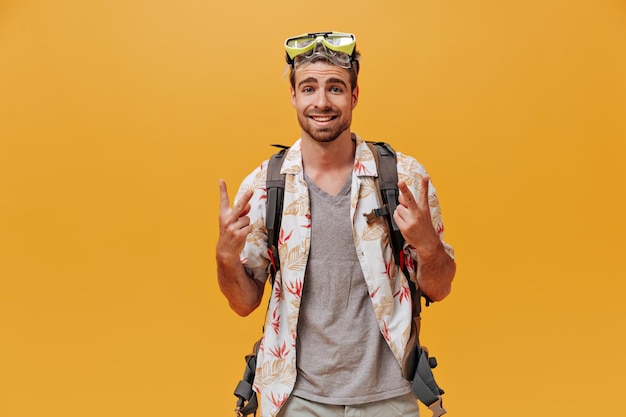 Foto gratuita hombre fresco con estilo con barba de jengibre y ojos azules en gafas de natación verdes y camisa blanca impresa que muestra signos de paz sobre fondo naranja