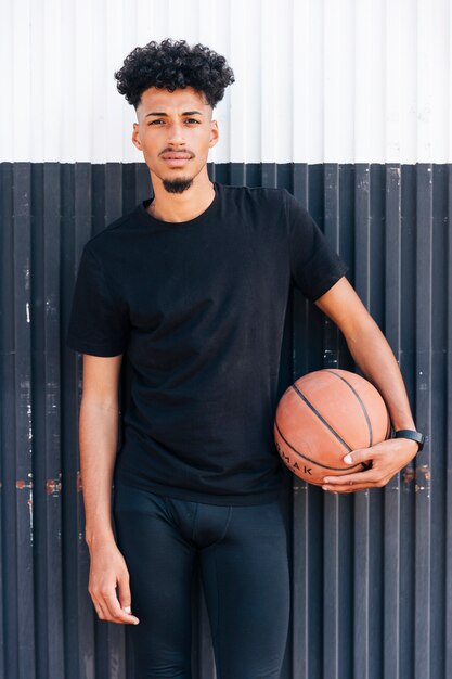 Hombre fresco joven que está parado contra la pared que sostiene baloncesto