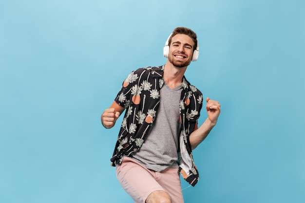 Foto gratuita hombre fresco de moda con barba en camiseta gris piña y camisa con estampado de palma y pantalones cortos sonriendo mirando a la cámara y posando con auriculares