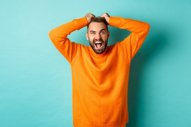 Foto gratuita hombre frustrado gritando, arrancándose el cabello y gritando enojado, perdiendo los estribos y luciendo enojado, parado sobre una pared turquesa clara.