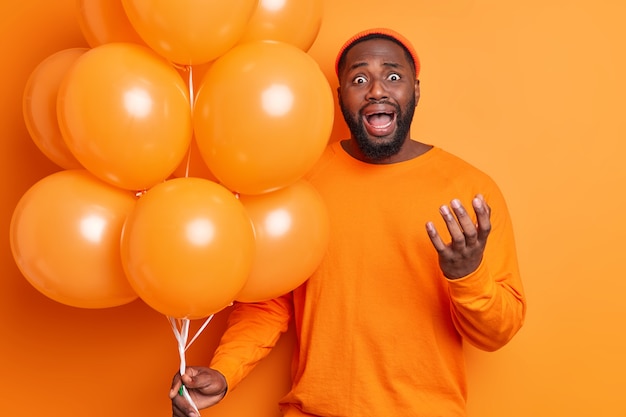 Hombre frustrado llora de desesperación exclama en voz alta gestos sostiene activamente un montón de globos de helio inflados usa sombrero y un jersey casual aislado sobre una pared naranja tiene vacaciones