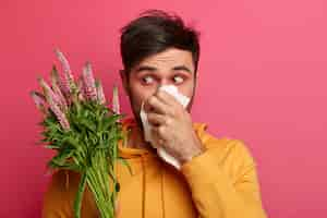 Foto gratuita el hombre frustrado se suena la nariz con un pañuelo, tiene enrojecimiento alrededor de los ojos, síntomas de alergia, tiene un aspecto poco saludable, se concentra en la flor, sufre de rinitis, reacción alérgica. personas y enfermedad