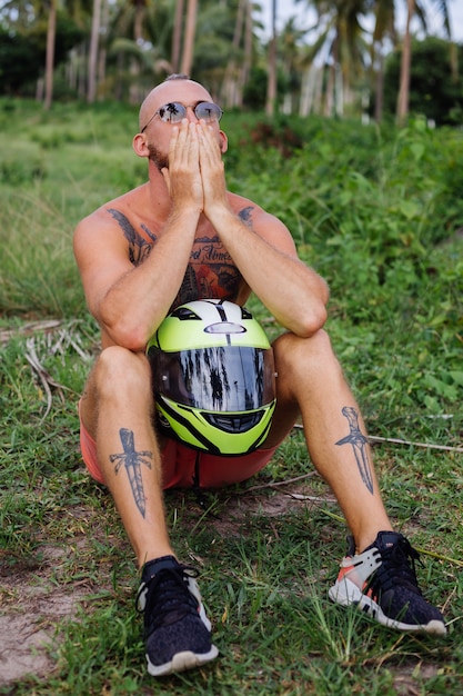 Hombre fuerte tatuado en el campo de la selva tropical con moto roja