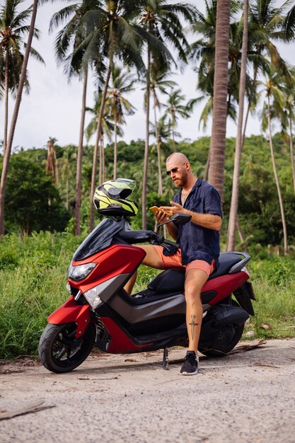 Hombre fuerte tatuado en el campo de la selva tropical con moto roja