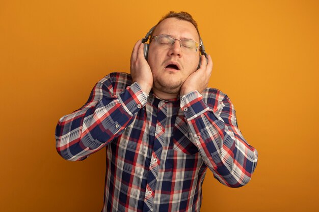 Hombre de gafas y camisa a cuadros con auriculares disfrutando de la música de pie sobre la pared naranja